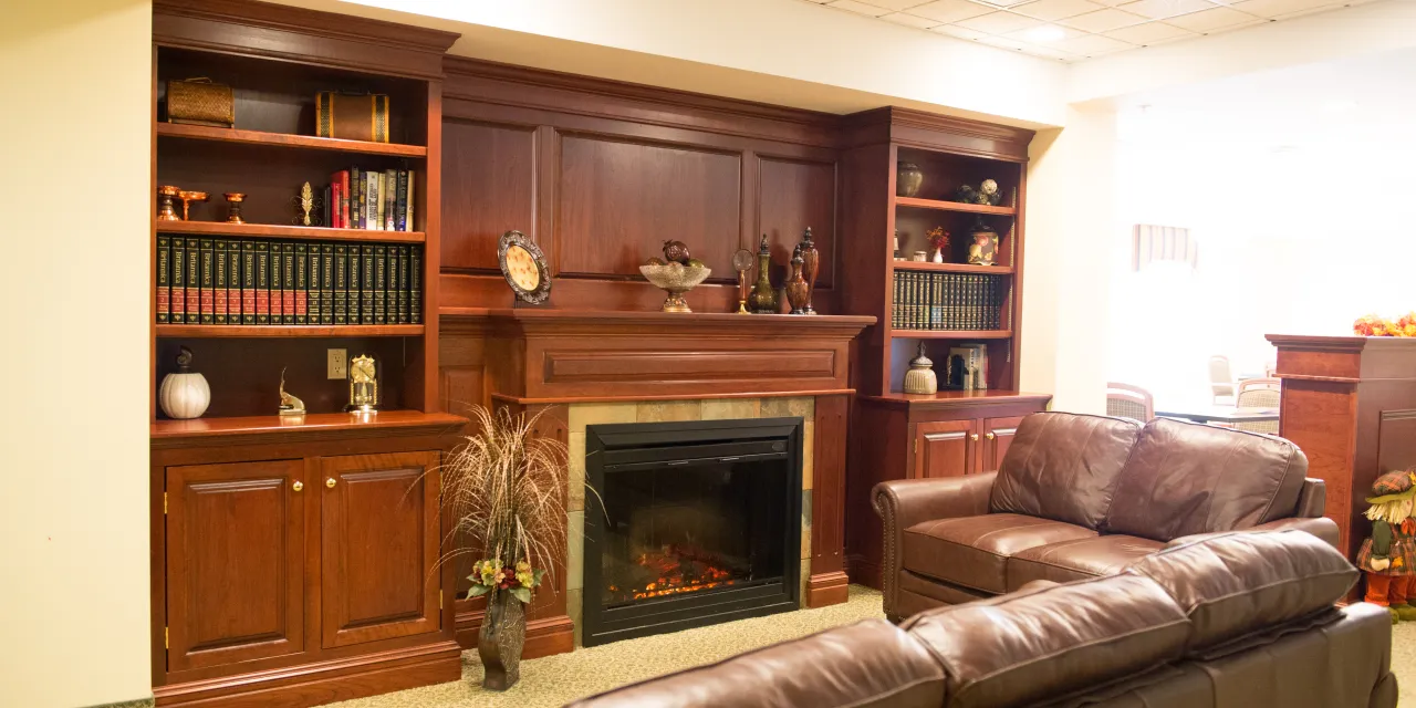 A grand fireplace with built in shelves surrounding it can be seen in a room with two large brown leather couches at the Memory Care Center
