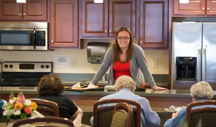 Residents in the Memory Care Dining facility sit and eat while a dietician stands at the counter smiling