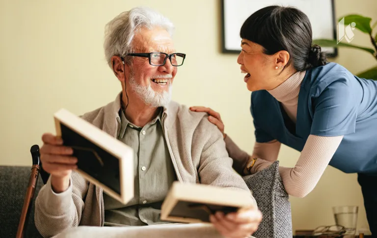 A nurse is looking at family photos along with an elderly male resident. His cane rests against the couch to his right, and he is smiling. 