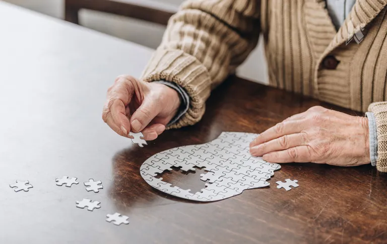 A senior man puts a puzzle together in the shape of a head. The missing pieces take up the space where the brain would be. 